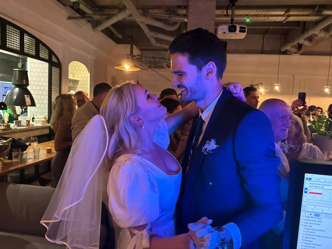 A couple is smiling at each other while dancing at a wedding, with the woman wearing a veil and the man in a suit. There are guests in the background.