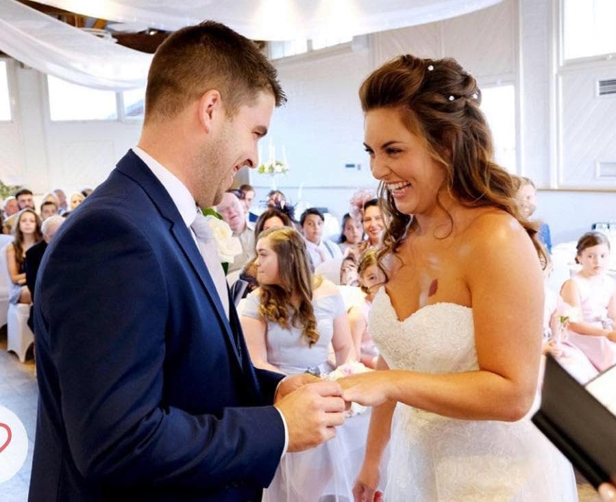 A bride and groom smiling at each other during a wedding ceremony, with the groom placing a ring on the bride's finger. Guests are seated in the background.