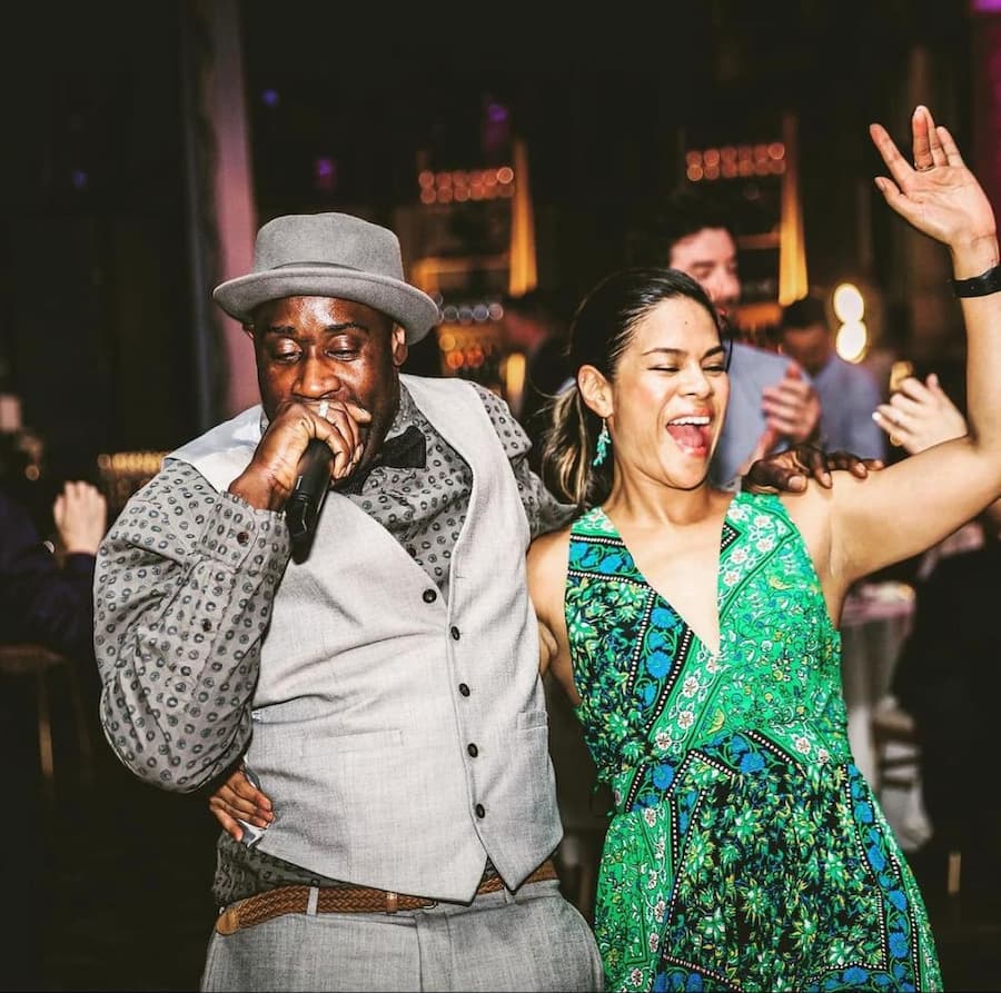 A man in a grey suit and hat sings into a microphone while dancing with a woman in a vibrant green dress, both enjoying a lively atmosphere.