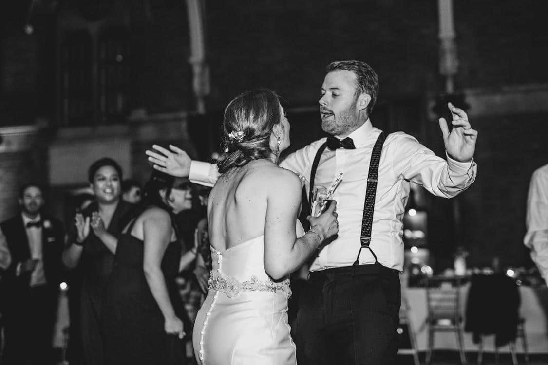 A joyful scene of a couple dancing at a celebration, with guests in the background clapping and enjoying the moment. The image is in black and white.