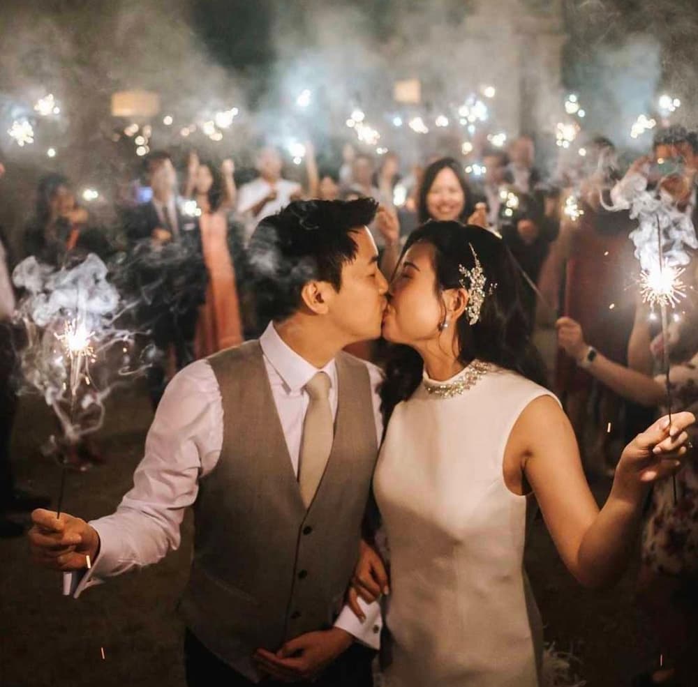 A couple is kissing while holding sparklers at their wedding, with guests celebrating in the background amidst smoke and twinkling lights.