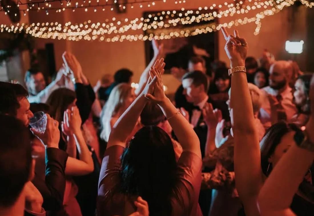 A lively gathering with people dancing and raising their hands, illuminated by string lights.
