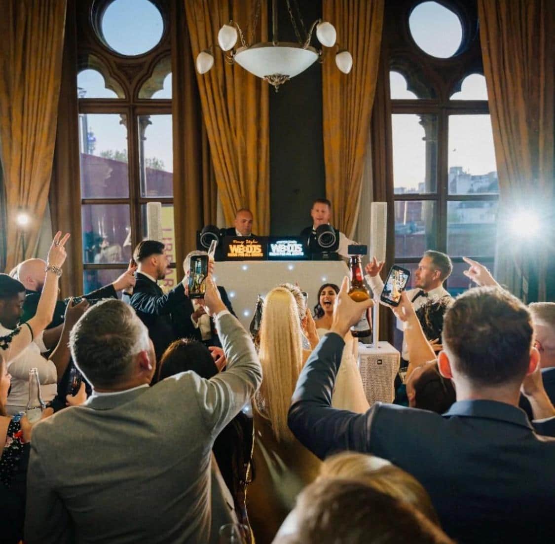 A lively celebration with guests raising drinks and using their phones, while a DJ performs in the background.