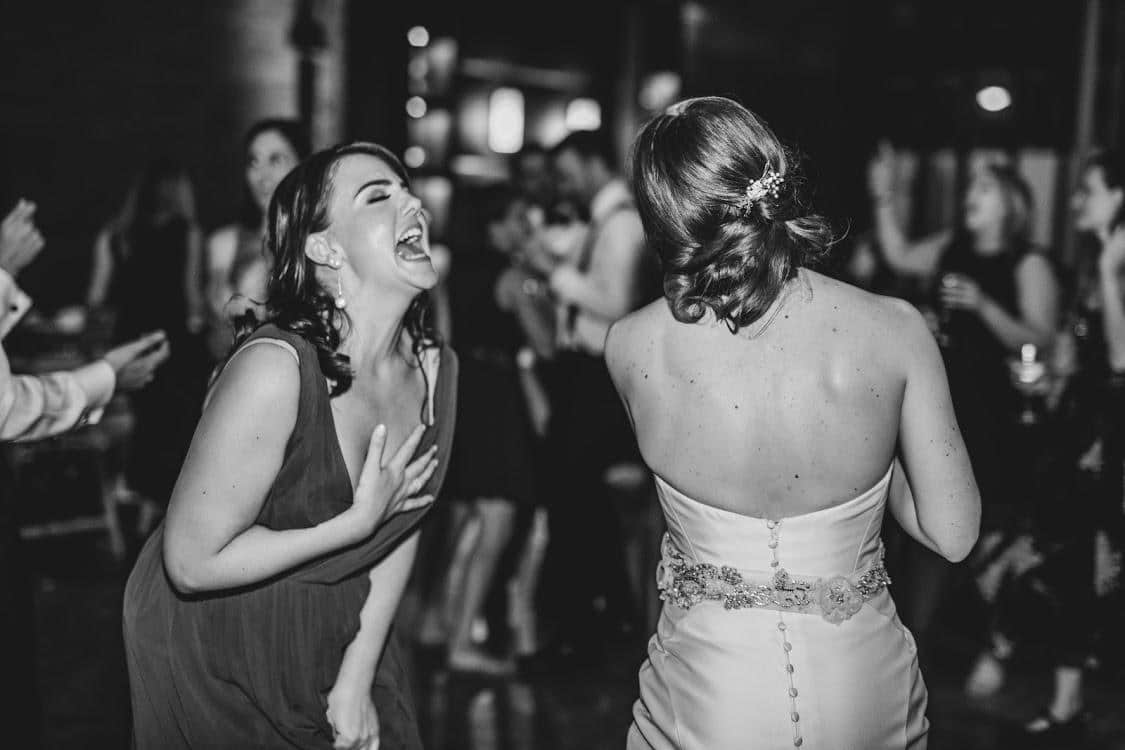 Two women are dancing and laughing at a celebration. One is wearing a dark dress and the other a wedding gown, with guests enjoying the background. The image is in black and white.