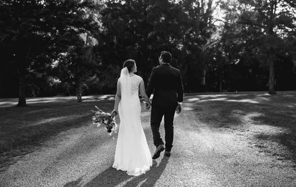A couple walking hand in hand down a gravel path, wearing wedding attire. The scene is in black and white, surrounded by trees.