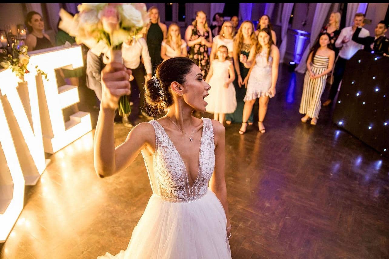 A bride in a white wedding dress holds a bouquet, celebrating with guests in a festive setting, illuminated by decorative lights spelling 'LOVE'.