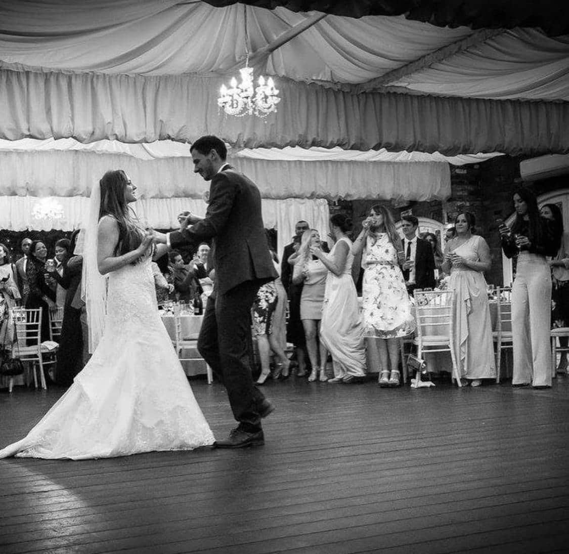 A bride and groom dance together at their wedding reception, surrounded by guests who are watching and celebrating. The setting features a decorated indoor area with a chandelier.
