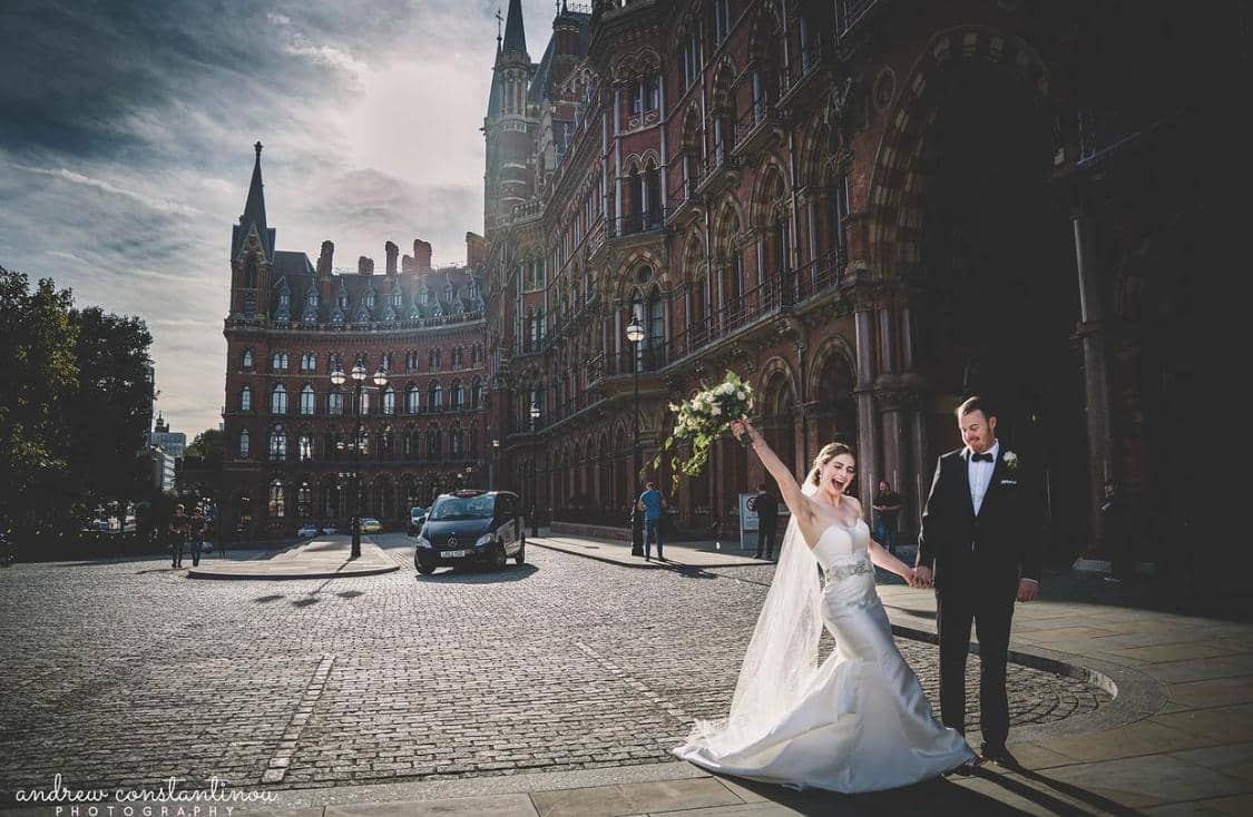 A joyful bride in a white wedding gown and a groom in a tuxedo hold hands outside a historic building, with the bride raising her bouquet in celebration.