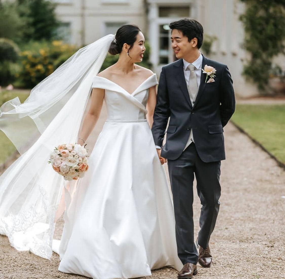 A bride in a white gown and a groom in a dark suit are walking hand in hand, smiling at each other, with a bouquet of flowers in the bride's hand. The setting is outdoors with greenery in the background.