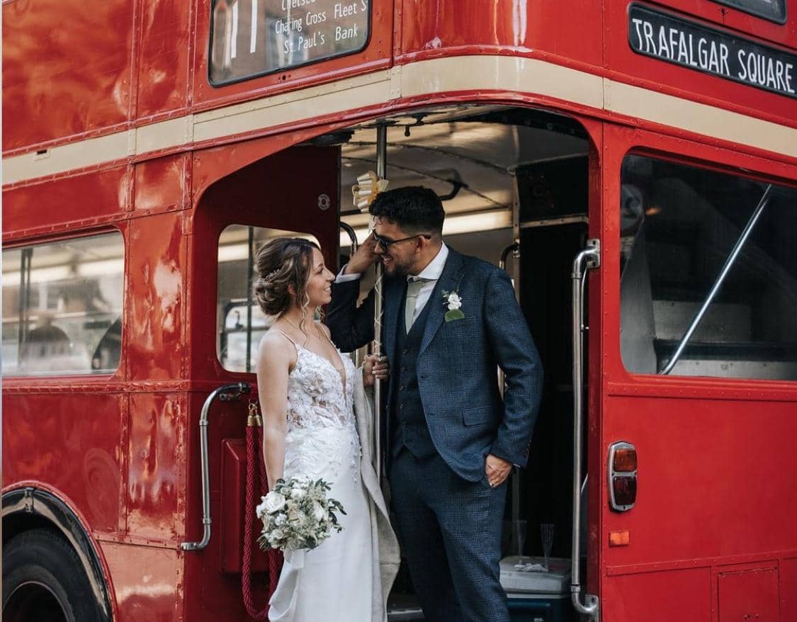 A bride and groom share a moment on the steps of a classic red double-decker bus. The bride holds a bouquet and wears a lace wedding dress, while the groom is in a blue suit with glasses.