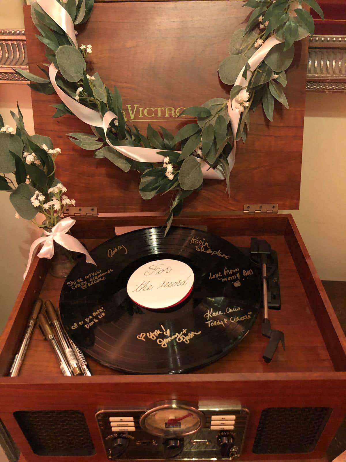 A vintage record player with a vinyl record inside, featuring handwritten messages on the record. It has a decorative wreath made of greenery and ribbons.