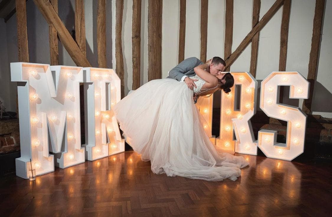 A newlywed couple sharing a kiss in front of large, illuminated letters spelling 'MR & MRS'. The bride is wearing a white gown, and the setting has a rustic backdrop.