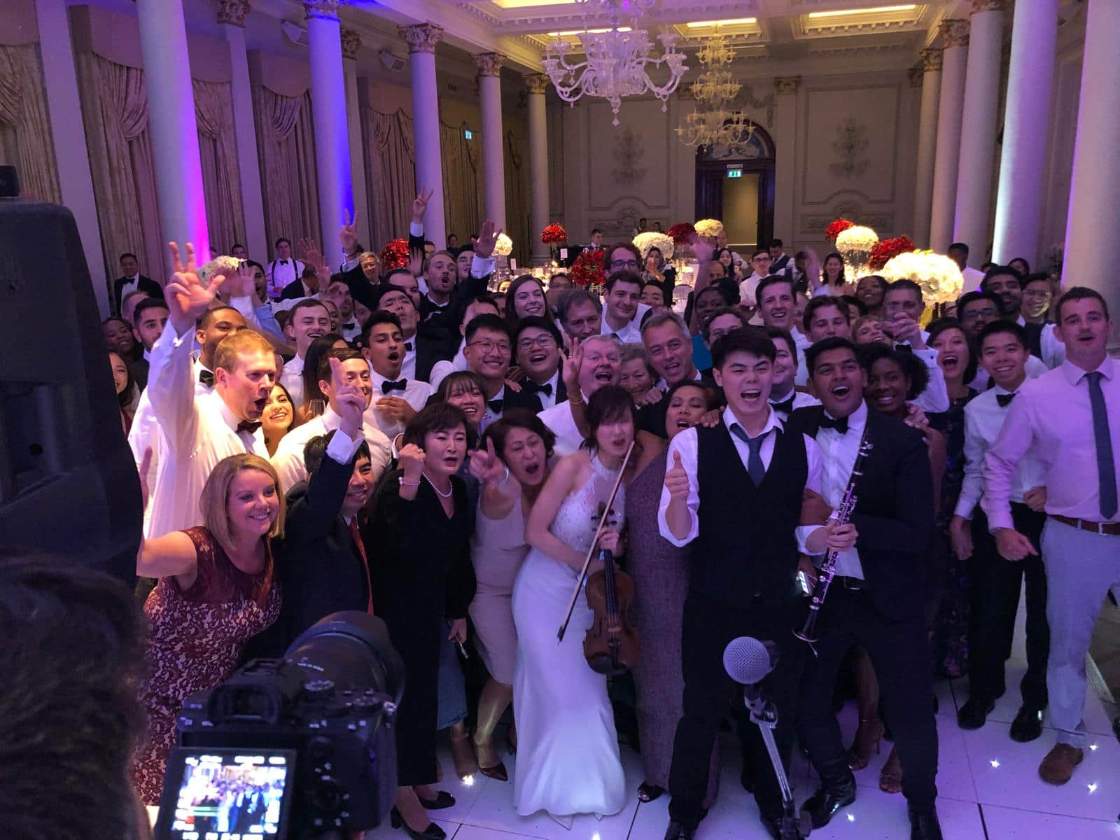 A large group of people joyfully posing together at a celebration, with a mix of formal attire and a performer holding a musical instrument.