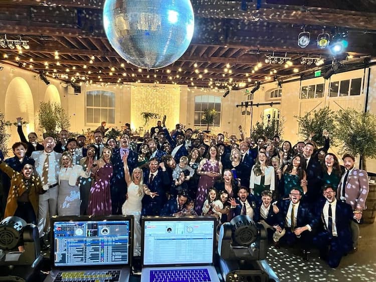 A large group of people celebrating at a festive event, with a disco ball overhead and party decor in a well-lit venue.