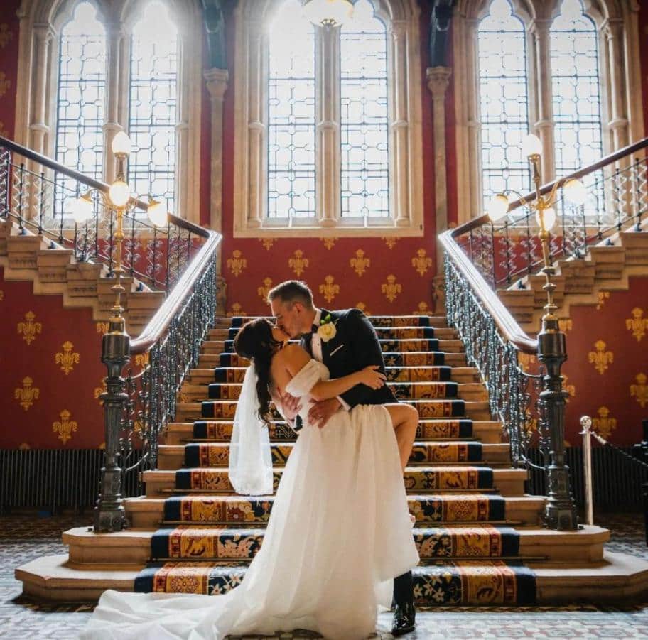 A bride and groom share a kiss on a grand staircase with ornate railings and stained glass windows in the background.