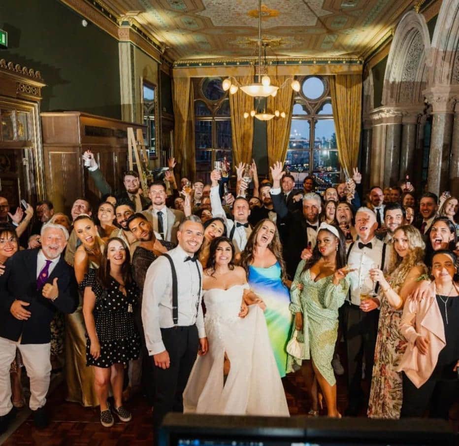 A large group of people celebrating at a wedding or event, with many smiling, posing, and raising drinks in a beautifully decorated indoor venue.