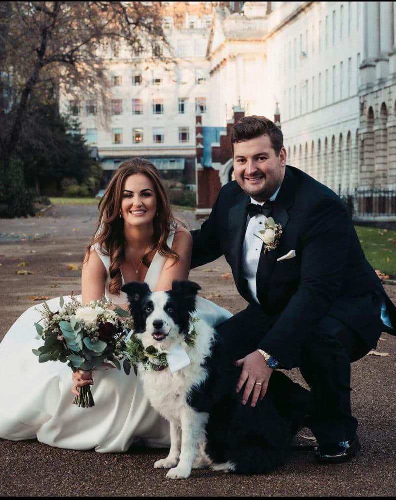 A couple in formal wedding attire poses together outdoors, holding a bouquet and accompanied by a dog wearing a floral collar.