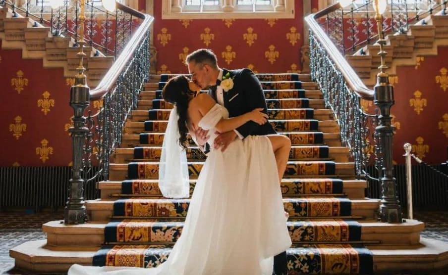 A romantic photo of a couple kissing on a grand staircase, decorated with ornate railings and a red patterned wall. The bride is in a white dress, and the groom is in a black suit with a boutonniere.