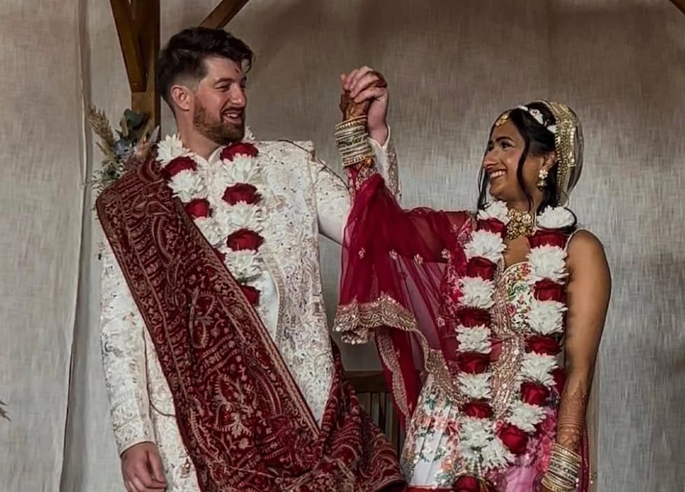 A man and woman are smiling at each other while holding hands, dressed in traditional wedding attire with floral garlands around their necks.
