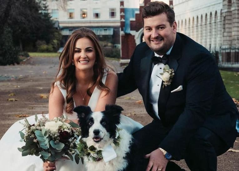 A bride and groom pose together with a dog, both holding flower bouquets, outdoors in a park setting.