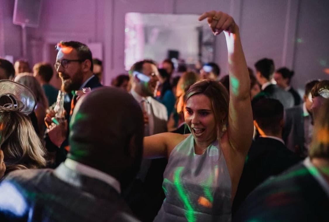 A lively party scene with people dancing and socializing, colorful lights reflecting in the background. A woman in a light dress raises her arm energetically.