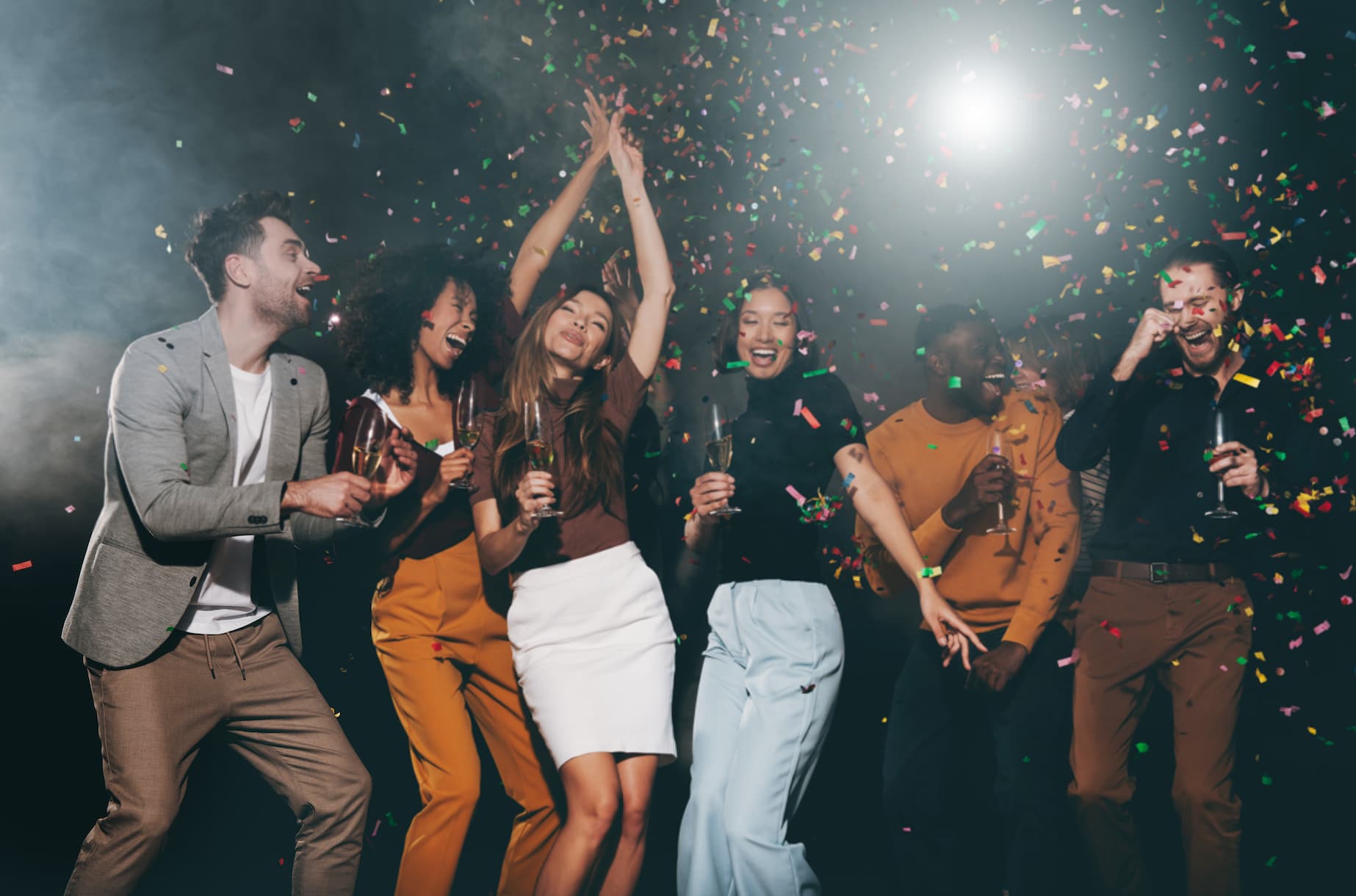 A group of people celebrating at a party, dancing and smiling, with confetti falling around them and holding drinks.