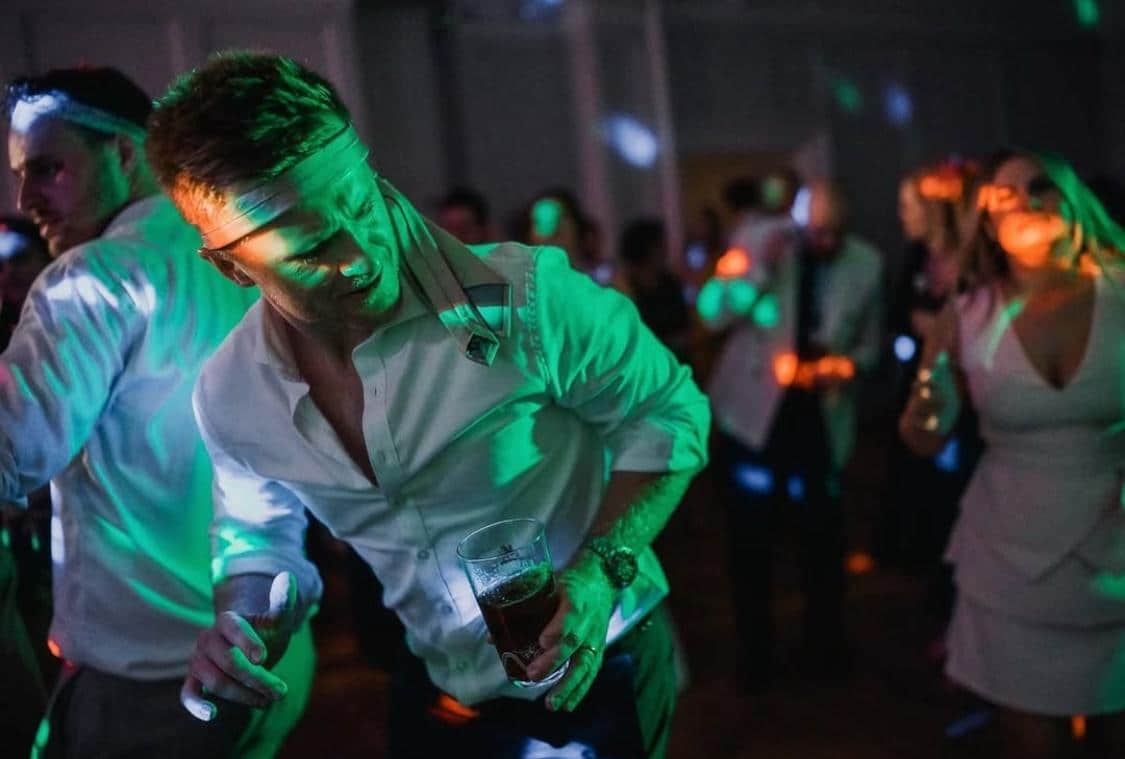 A man in a white shirt dances at a party while holding a drink, surrounded by people and colorful lights.