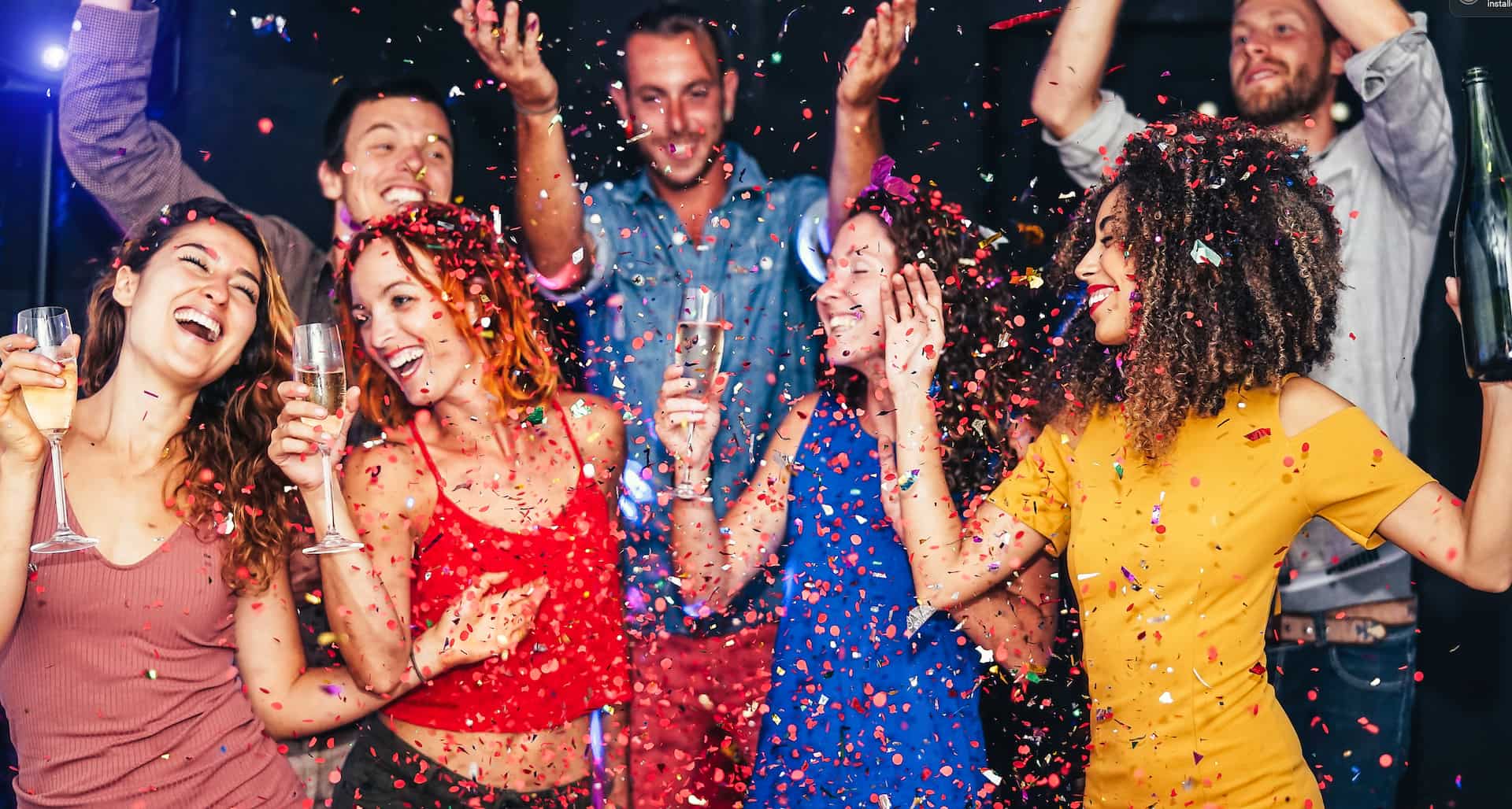 A group of people celebrating at a party, smiling and throwing confetti, with drinks in hand.