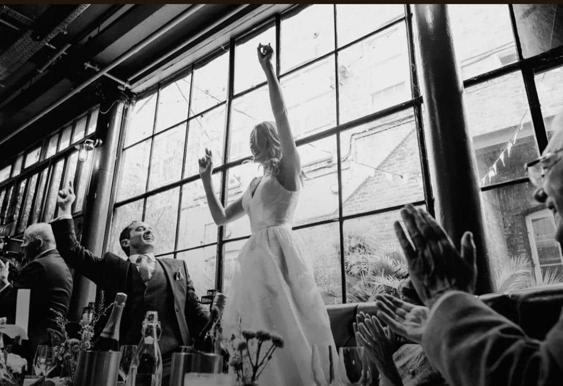A woman in a white wedding dress is dancing joyfully, with arms raised, while others around her cheer and applaud. The setting features large windows and festive decorations.