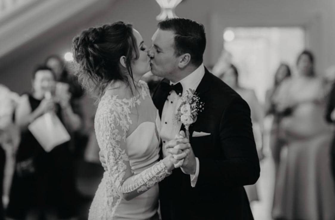 A bride and groom are sharing a kiss while holding hands, surrounded by guests celebrating in the background. The image is in black and white.