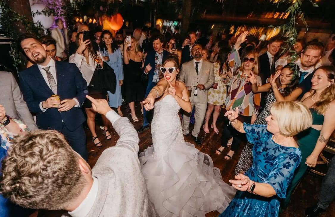 A bride in a white wedding dress dances energetically in a lively crowd at a celebration, surrounded by guests enjoying the moment.