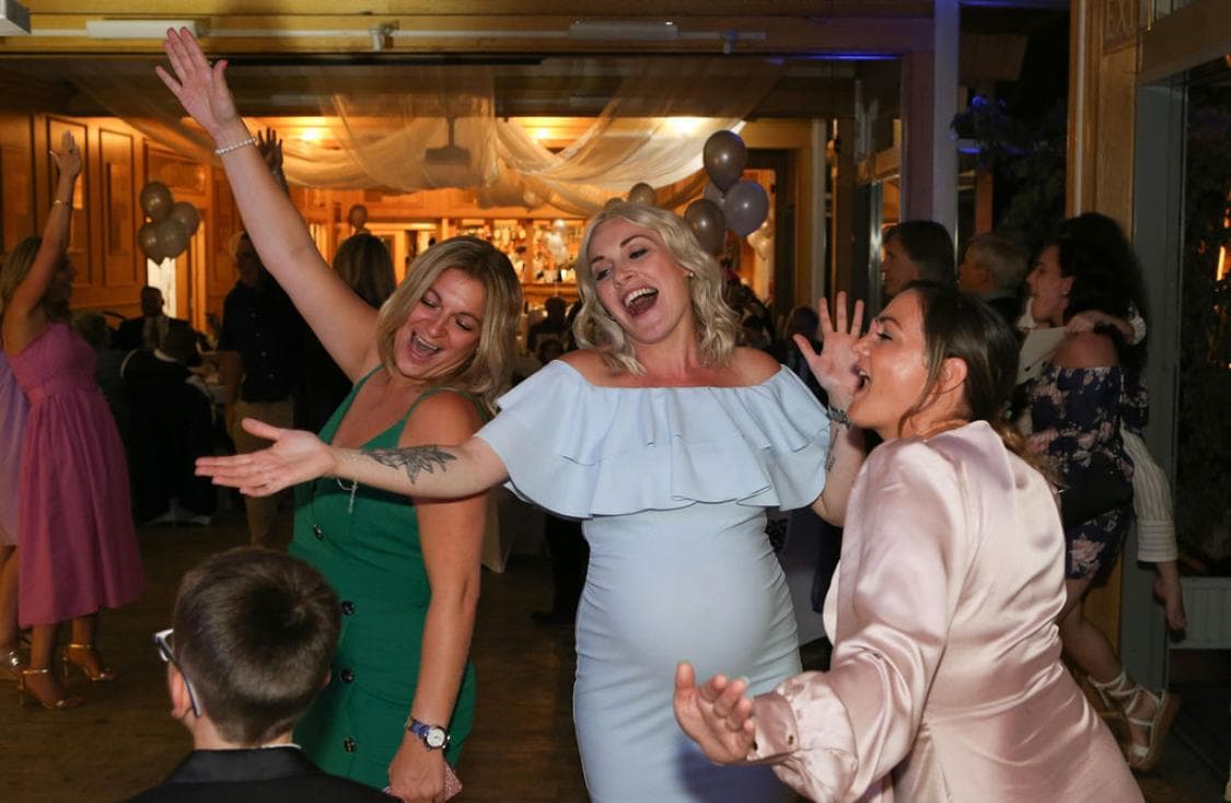 A group of three women dancing and enjoying themselves at a celebration, with balloons and guests in the background.