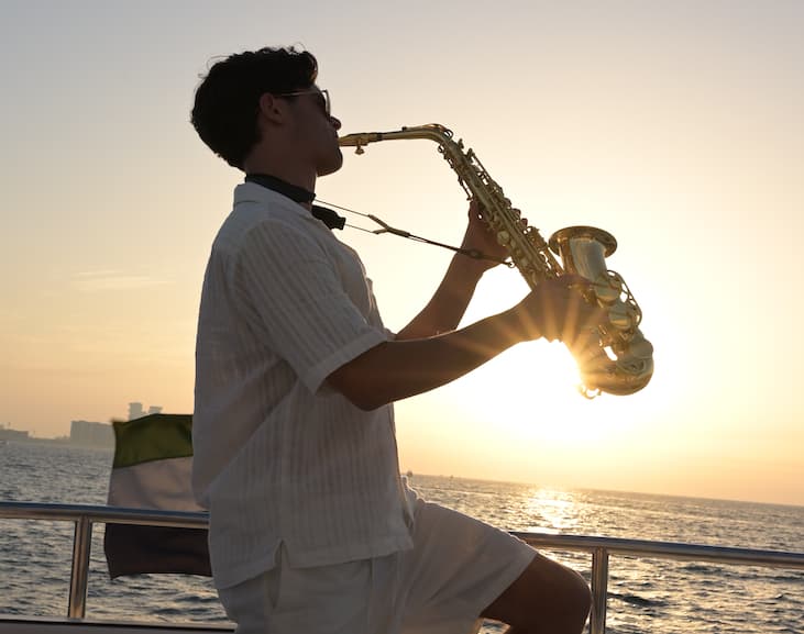 A young man playing a saxophone on a boat during sunset.