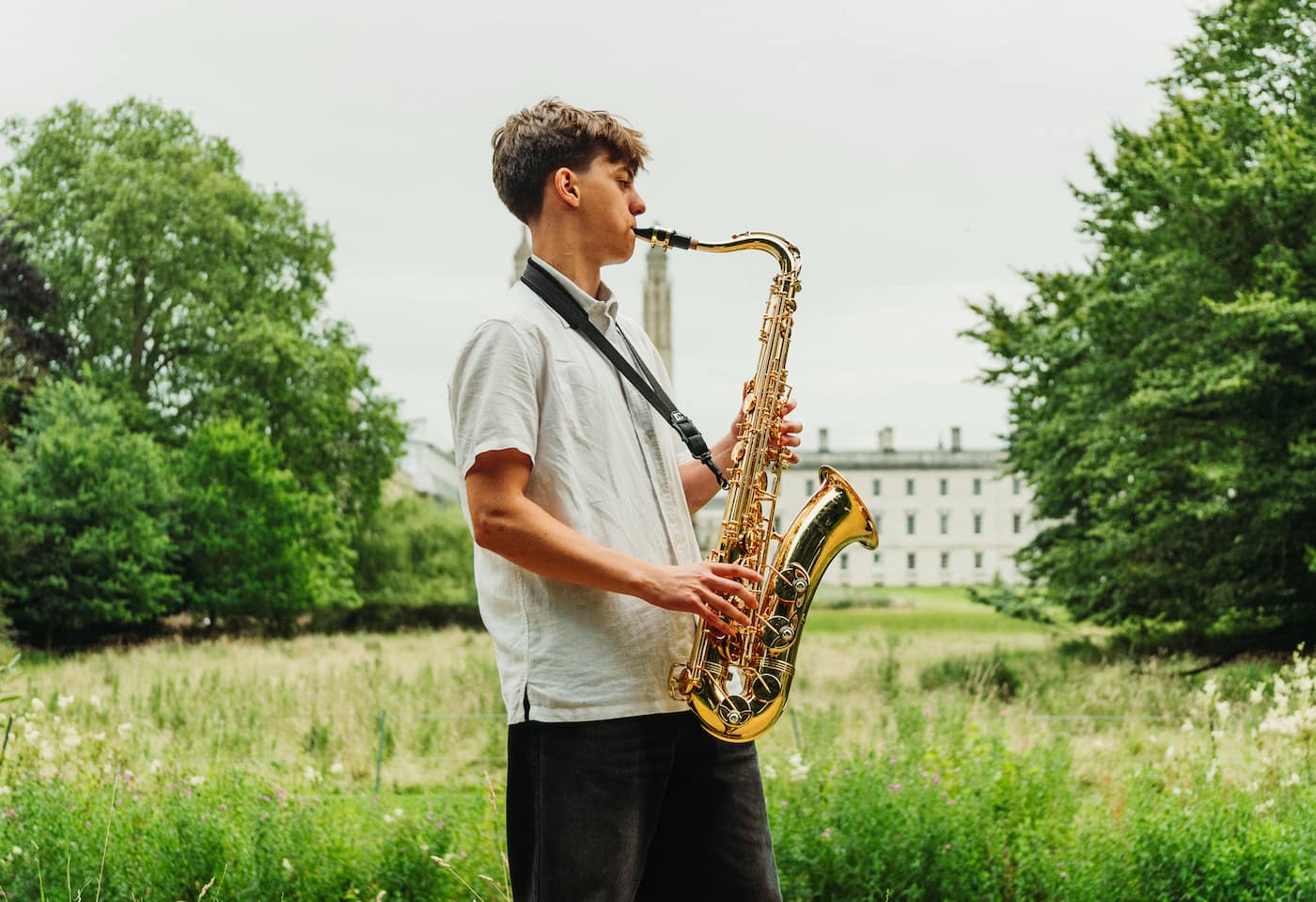 A young man playing a saxophone in a green outdoor setting with buildings in the background.