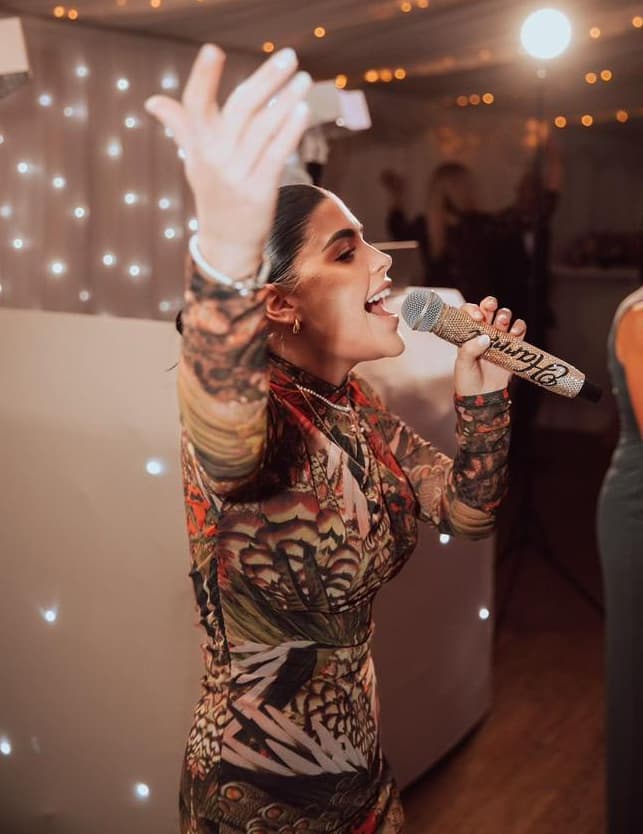 A woman is singing into a microphone while wearing a colorful, patterned dress. She appears joyful and engaged in a lively atmosphere with soft lights in the background.