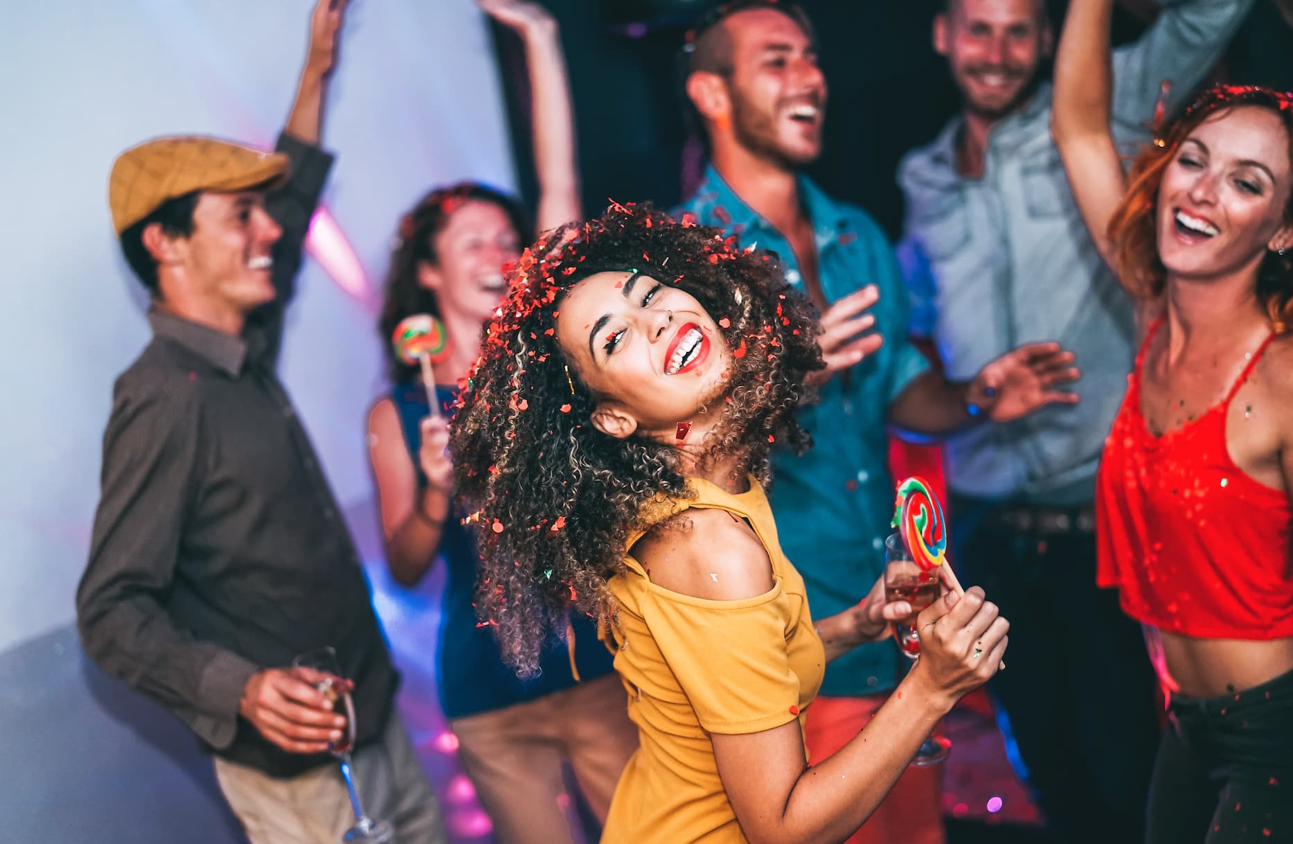 A lively group of people celebrating at a party, with colorful confetti, drinks, and a cheerful atmosphere. A woman in a yellow top is smiling and dancing, holding a lollipop.