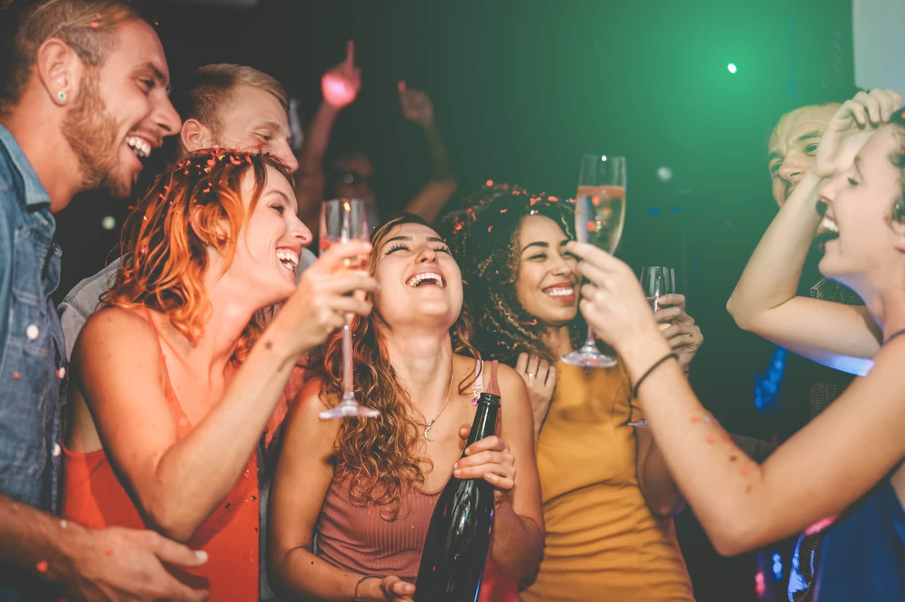 A group of friends celebrating and laughing together at a party, holding drinks, with confetti in the air.