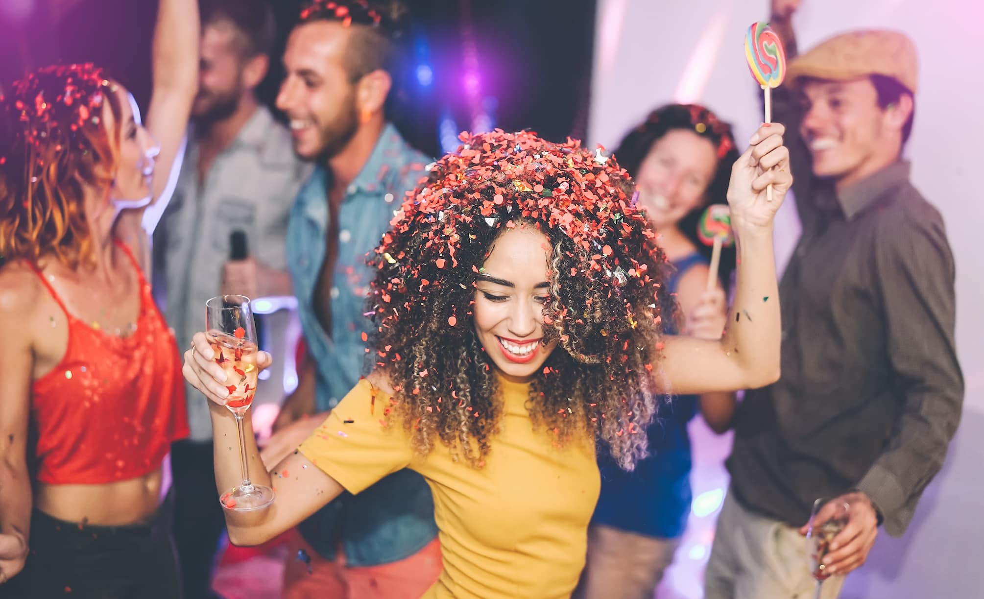 A group of people celebrating at a party, with one woman dancing joyfully, wearing a yellow shirt and adorned with colorful confetti in her hair. Others are holding drinks and lollipops, and the atmosphere is vibrant and festive.