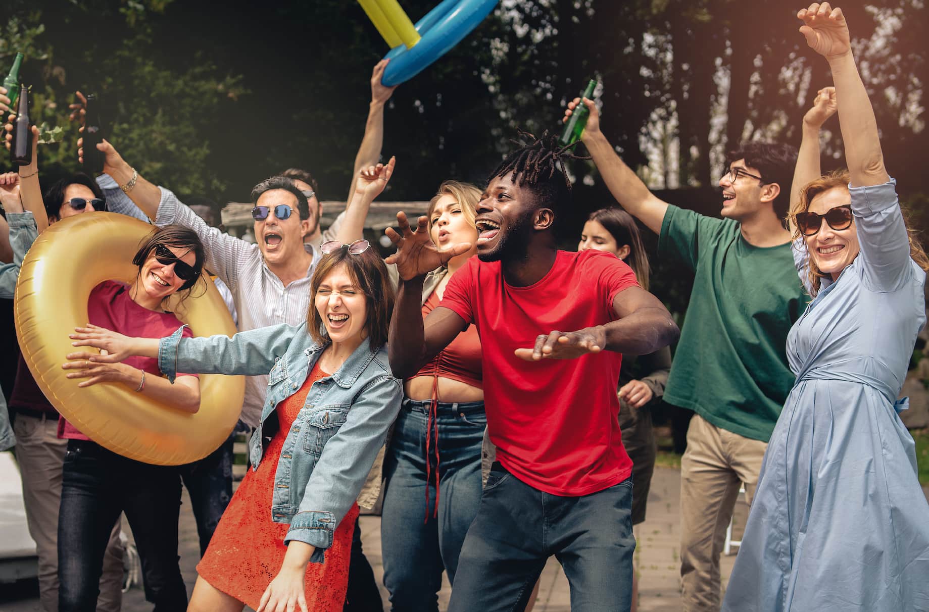 A group of joyful people celebrating outdoors, dancing and holding drinks, with some wearing sunglasses and colorful attire.