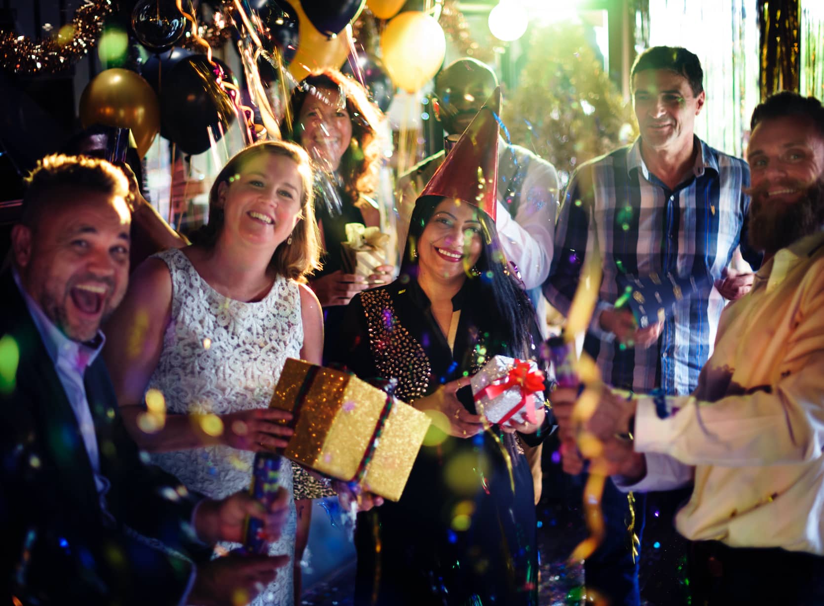A festive group of people celebrating, smiling, and holding gifts with balloons and decorations in the background.