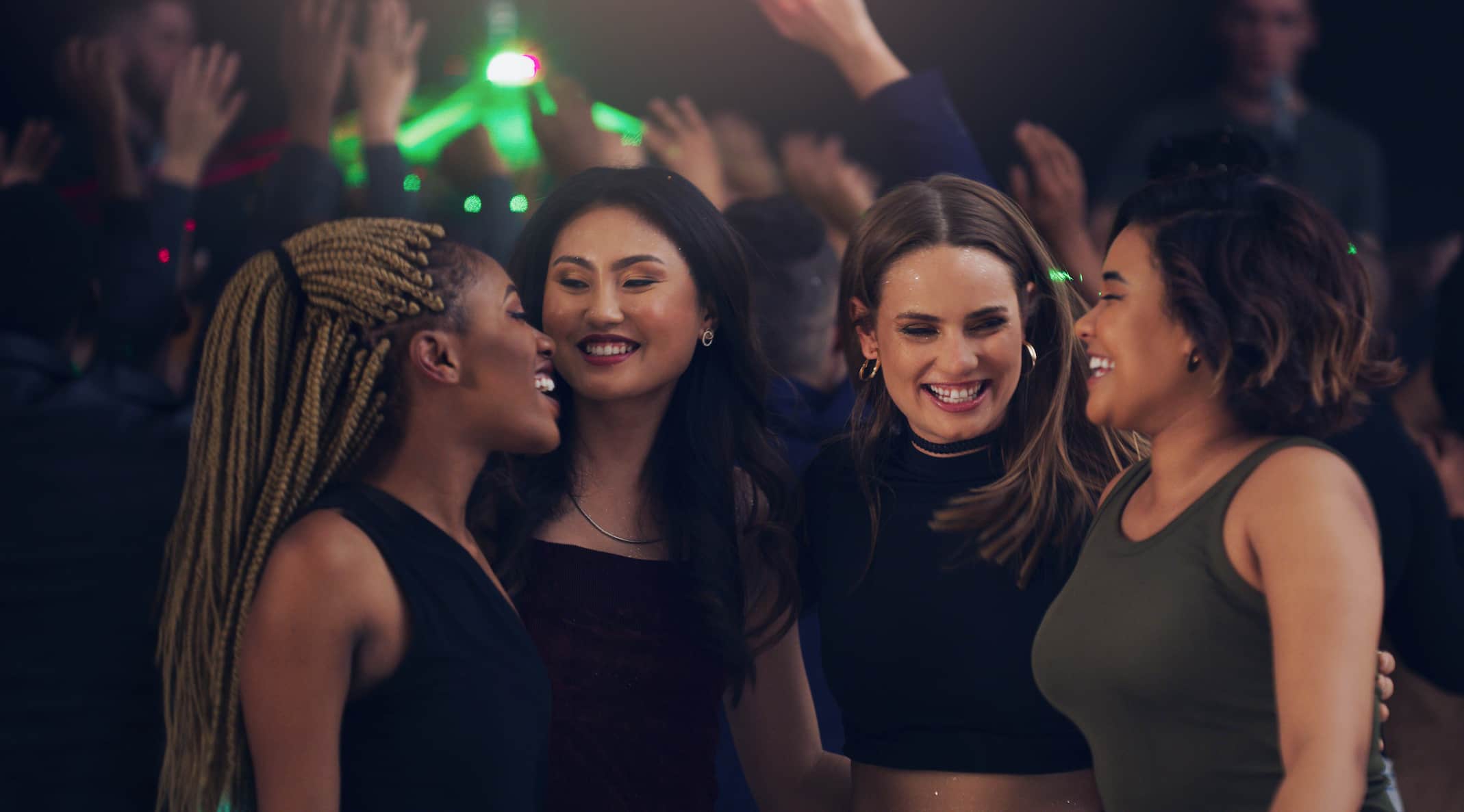 Four women are smiling and enjoying each other's company at a lively party, with hands raised and lights in the background.