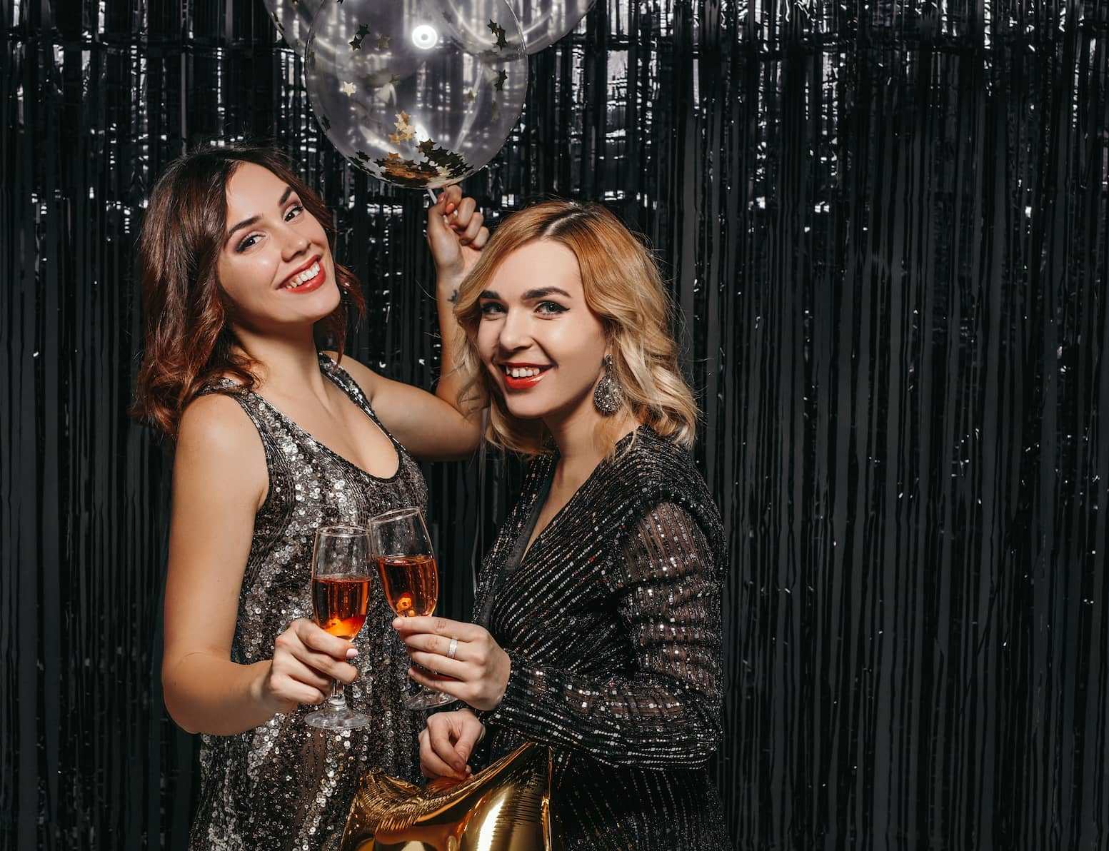 Two women in sparkling dresses hold drinks and smile, celebrating at a party with a shiny black backdrop and a balloon.