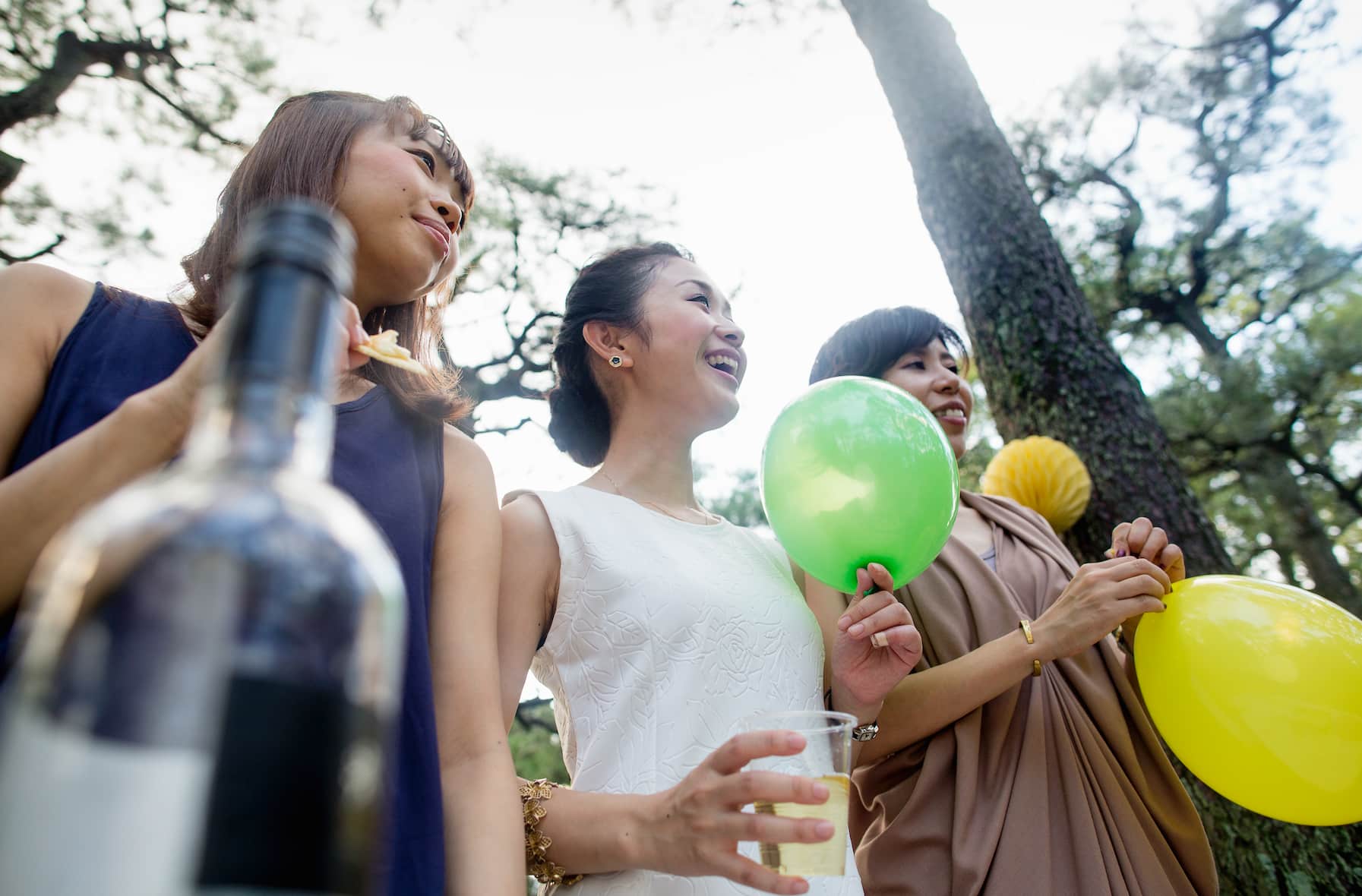 Three women are smiling outdoors, holding balloons and drinks, with trees in the background.