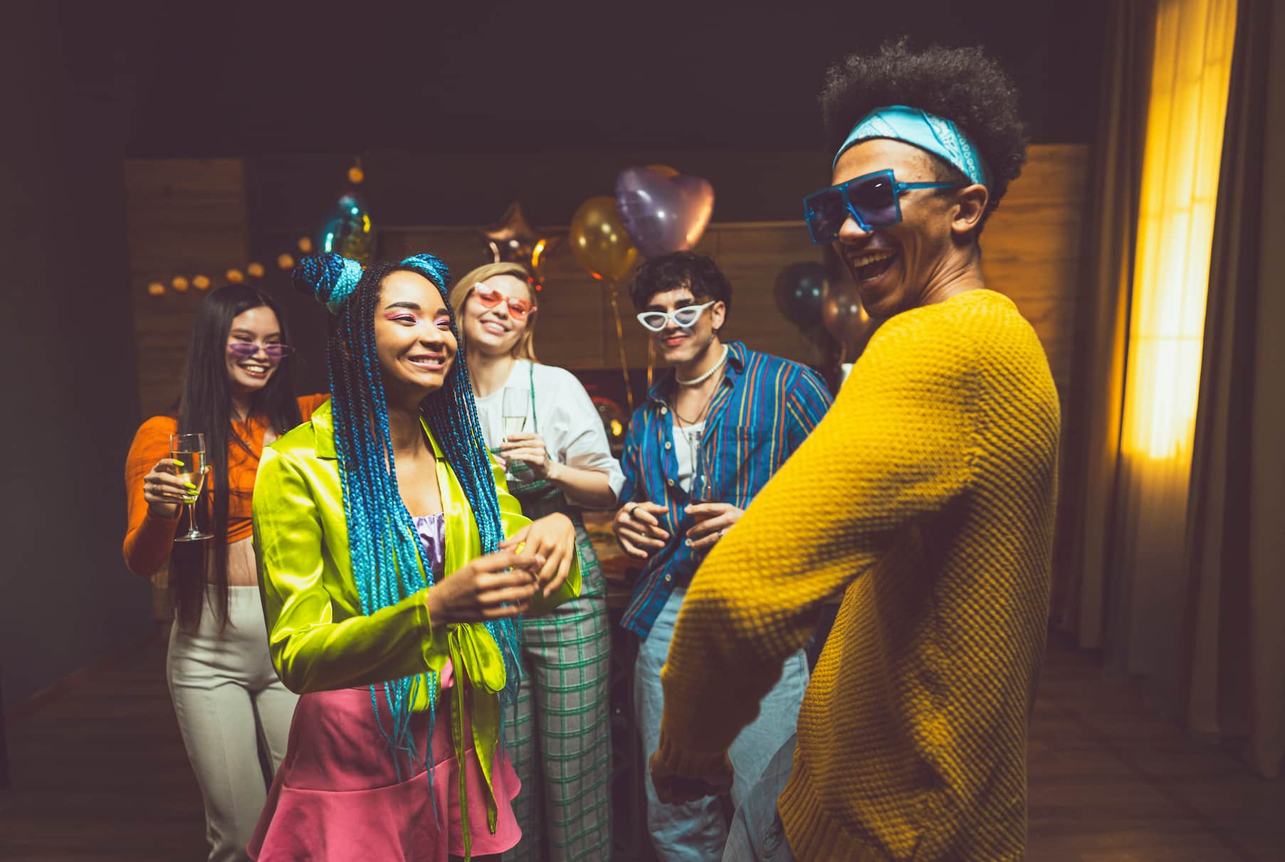 A group of five young people celebrating at a party, wearing colorful outfits and sunglasses, holding drinks and smiling. Balloons are in the background.