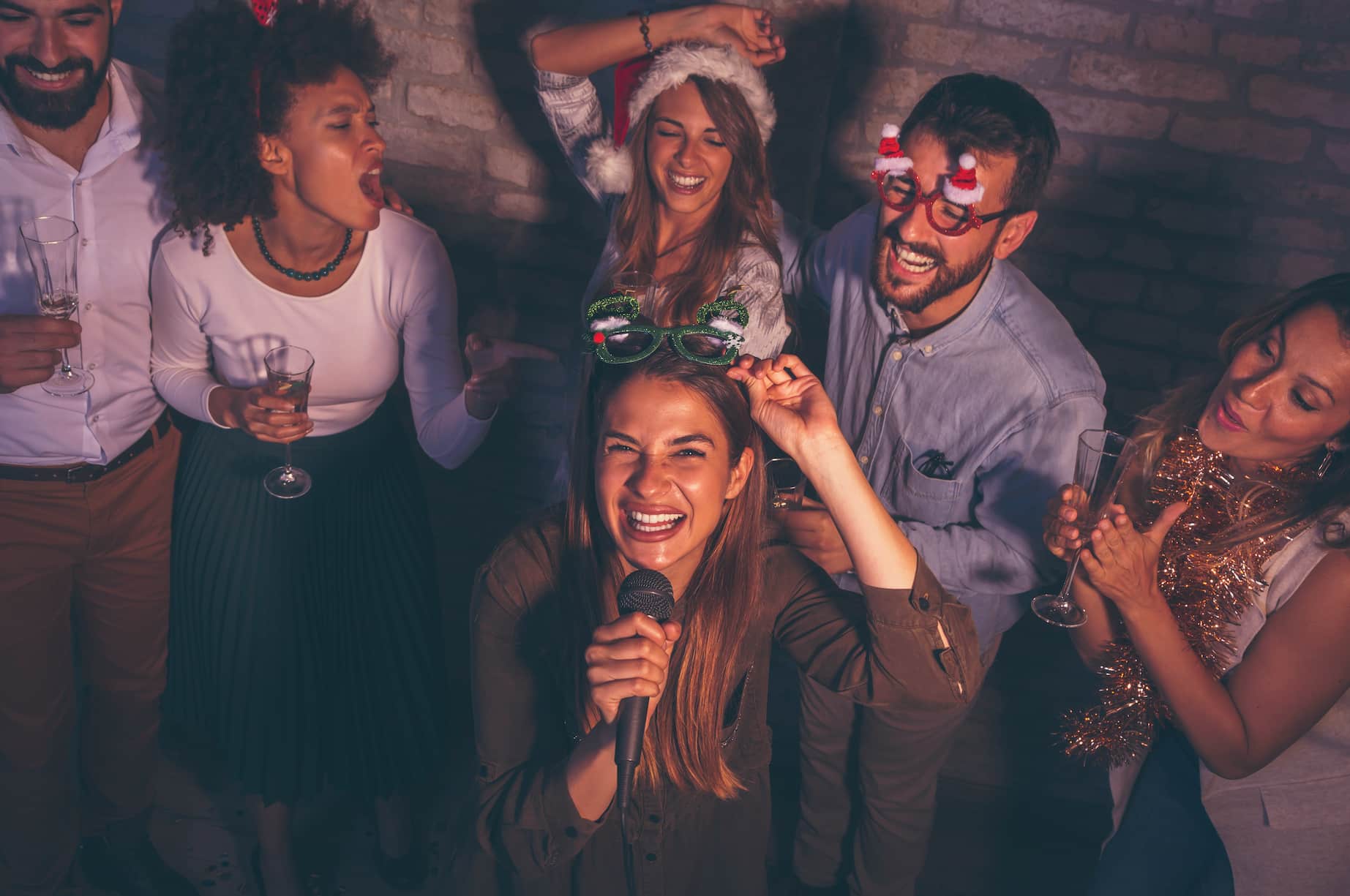 A group of friends celebrating joyfully at a party, some wearing festive accessories, with one person singing into a microphone.