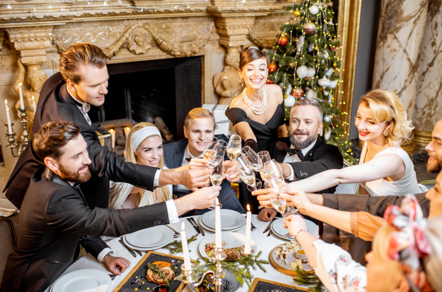 A group of elegantly dressed people are toasting with champagne glasses at a festive dinner table, decorated with food and candles, and a decorated Christmas tree in the background.