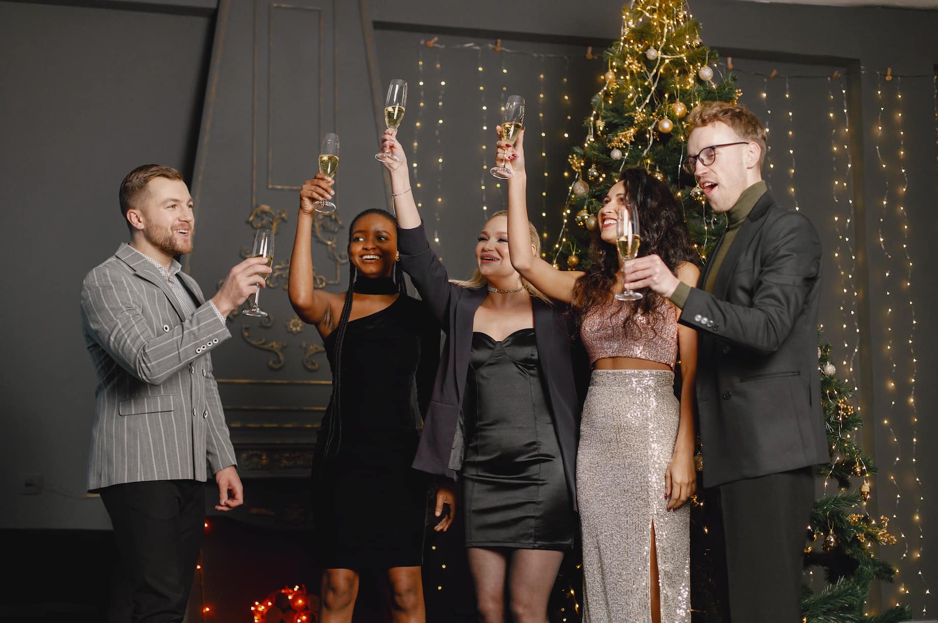 A group of five people, dressed in formal attire, raise their champagne glasses in celebration in front of a Christmas tree decorated with lights.