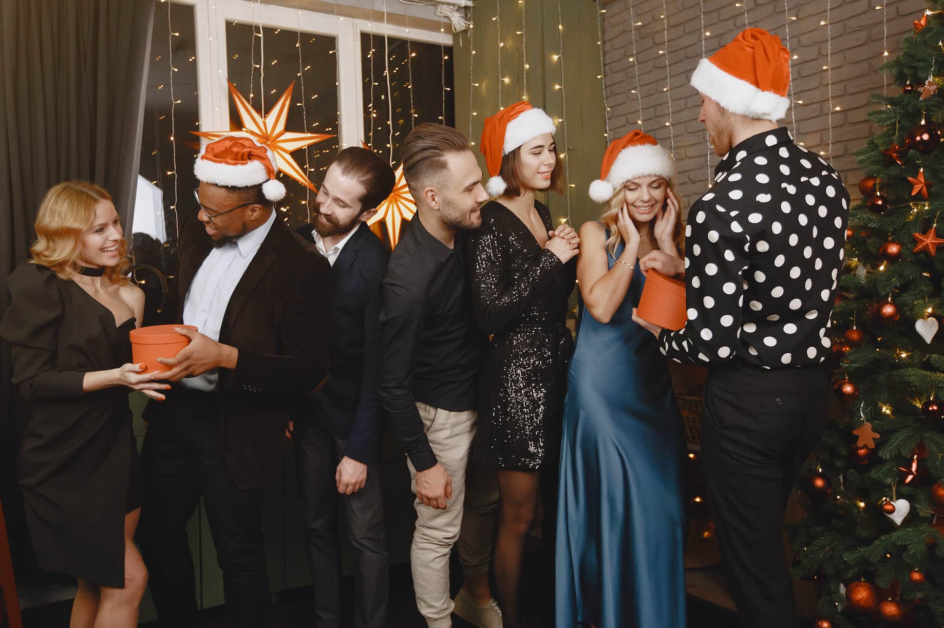 A group of six people wearing Santa hats, celebrating near a decorated Christmas tree, with one person holding a terracotta pot and smiling.