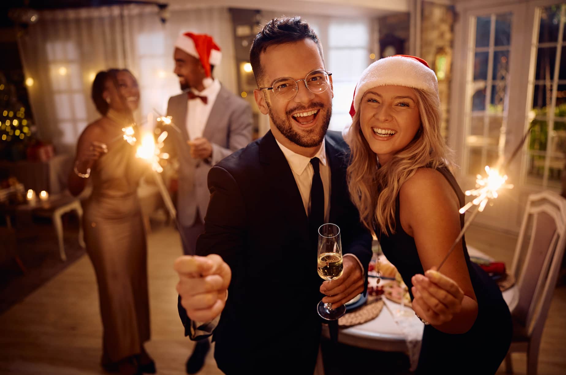 A group of four friends celebrates together in a festive setting. Two of them hold sparklers, while one has a champagne glass. The atmosphere is joyful, with holiday decorations and soft lighting.