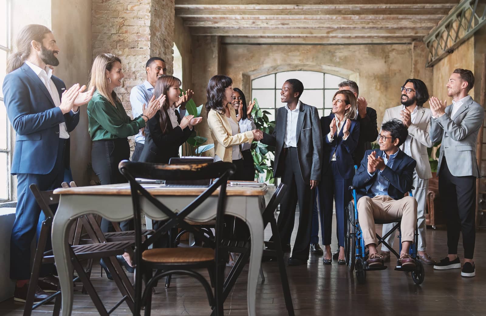 A group of professionals is applauding and celebrating, with two individuals shaking hands at the center. The setting is a modern office with exposed brick and large windows.